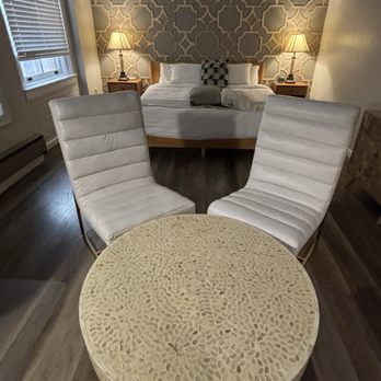 The Carrick - Seating area in master bedroom - The image shows a bedroom with a bed against a patterned accent wall, flanked by two nightstands with lamps. In the foreground, there are two white, padded chairs positioned around a round, textured table. The room has a window with blinds on the left side, and the flooring is dark wood. - Hotels Near Me - Bisbee, Arizona