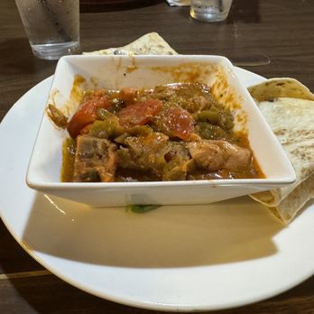 El Bruno's - Green Chile stew - A bowl of green chile stew containing pieces of meat, tomatoes, and peppers is placed on a white plate. The stew has a thick, flavorful broth. Two folded tortillas are positioned on the side of the plate. The setting appears to be a restaurant with a wooden table, and two glasses of water are visible in the background. - Mexican Near Me - Albuquerque, New Mexico