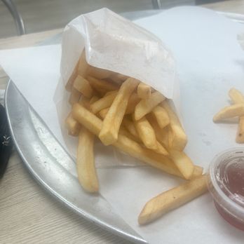 Original Geno's - French fries are placed in a paper wrapper on a metal tray, with a small container of ketchup nearby, on a light-colored table. - Pizza Near Me - Tempe, Arizona