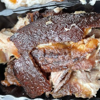 Taino's Kitchen - The image shows a close-up of cooked pork with crispy, browned skin and tender meat, served in a black container. The pork appears to be prepared in a traditional style, possibly for a Puerto Rican dish like lechón or pernil, with visible textures of crispy skin and moist meat. The background includes some other food items, possibly rice or side dishes, indicating a meal setting. - Puerto Rican Near Me - Newark, New Jersey