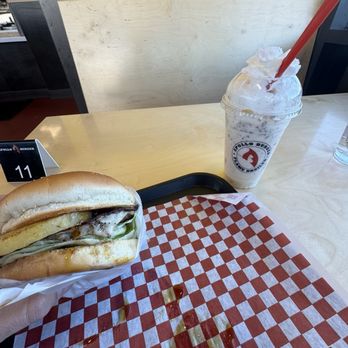 Apollo Burger - Hawaiian Teriyaki Burger and reeses shake - A burger with lettuce, cheese, and meat is held in a hand over a checkered paper on a table, with a milkshake in a clear cup nearby. - Burgers Near Me - Lehi, Utah