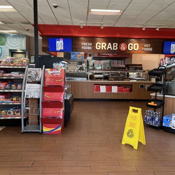 RaceTrac - a view of a grocery store - The image shows the interior of a convenience store with a grab & go food counter labeled 'FRESH FOODS GRAB & GO HOT FOODS.' To the left, there are snack displays including Skittles Littles and other snack items. On the right, bottled water and oranges are visible on a small black shelf. A yellow caution sign is placed on the wooden floor in front of the counter. The ceiling has white tiles with fluorescent lighting, and digital screens are mounted above the counter. - Gas Stations Near Me - St Augustine, Florida