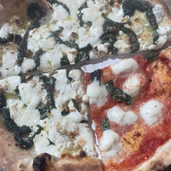 Pupatella - Pesto Pizza and Margherita DOC Pizza. - The image shows two pizza slices on a white plate. The first slice has pesto sauce, cheese, and chopped nuts, with a slightly charred crust. The second slice features tomato sauce, melted mozzarella cheese, and basil leaves, with a golden-brown crust. The plate is placed on a wooden surface, and part of a metal utensil is visible in the corner. - Pizza Near Me - West Springfield, Virginia