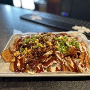 Wolfies Grill - Pendleton - Pork Nachos - A serving of pork nachos on a paper-lined tray at Wolfies Grill in Pendleton, Indiana. The nachos include tortilla chips topped with shredded pork, barbecue sauce, chopped green onions, and corn kernels. The background shows a bar area with a black rubber mat and some white objects, likely bar accessories or condiments. The setting appears to be inside a modern family grill restaurant with natural lighting. - Bars Near Me - Pendleton, Indiana
