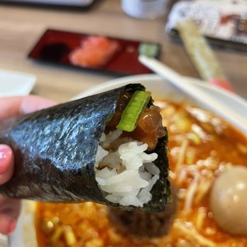 Chicago Sushi - Hand Roll Spicy Tuna - A person holds a spicy tuna hand roll with rice, seaweed, and a green onion topping. In the background, there is a bowl of ramen with broth, noodles, and toppings, along with a small tray of condiments and utensils on a wooden table. - Japanese Near Me - Des Plaines, Illinois