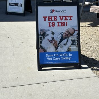 Tractor Supply - Vet clinic - A portable sidewalk sign outside PetVet features the message 'The Vet Is In!' along with an image of a veterinarian in a white coat with glasses, holding a dog. The sign promotes walk-in veterinary care and is positioned on a concrete sidewalk near outdoor seating and other signage. The background includes a stone wall and outdoor furniture, indicating a commercial or retail area. - Department Stores Near Me - Apache Junction, Arizona