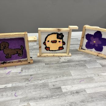 Tuft A Rug - Three tufted art pieces with wooden frames are displayed on a gray floor, depicting a dog, a chick, and a purple flower. - Art Classes Near Me - Aurora, Illinois