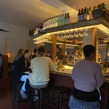 The Fly - a group of people sitting at a bar - The image shows a bar area inside a restaurant, with several patrons seated on bar stools. The bar features a display of bottles and glasses on overhead shelves, and the background includes bartenders preparing drinks. The lighting is warm, and the setting appears lively with multiple people engaged in conversation. - Wine Bars Near Me - Brooklyn, New York