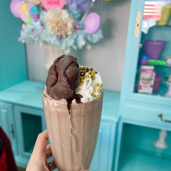 Kdulche Cafe - Nutella Shake - A tall glass of Nutella milkshake with whipped cream, chocolate drizzle, and gold sprinkles, held in front of a colorful cafe background. - Coffee & Tea Near Me - Lansing, Illinois