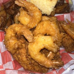 Jumbo Shrimp - The image shows a serving of fried shrimp with a golden, crispy coating, accompanied by seasoned, cooked potatoes and a piece of cornbread. The food is arranged on a checkered paper lining, indicating a takeout presentation from a restaurant specializing in fried seafood and chicken dishes.