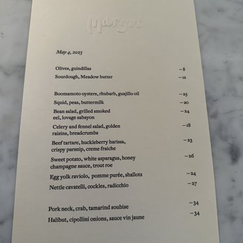 Margot - Menu - A vertically oriented menu with embossed Margot logo at the top, listing dishes and prices, set on a marble table. - New American Near Me - Brooklyn, New York