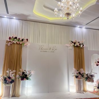 TDE Wedding - Welcome back-drop - The image shows a wedding backdrop setup in an event hall with white drapery and floral arrangements in pink, white, and dark purple. The backdrop includes a white panel with the names Theresa & Kai and the date December 6, 2025. The ceiling has a decorative chandelier and recessed lighting, and the floor is covered with a patterned carpet. The overall scene is decorated for a wedding ceremony or reception. - Wedding Planning Near Me - Millbrae, California