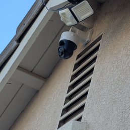 Two security cameras and a motion sensor light are mounted on an exterior wall of a building, with a vent and part of the roof visible.