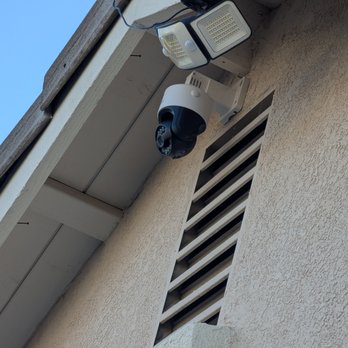 On Time Solutions - Two security cameras and a motion sensor light are mounted on an exterior wall of a building, with a vent and part of the roof visible. - Security Systems Near Me - Temecula, California