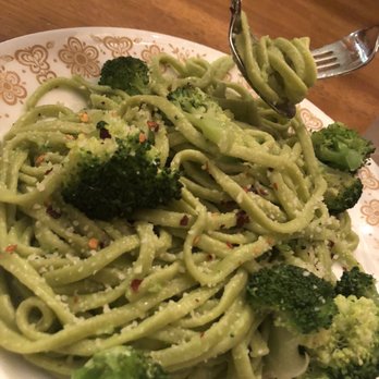 Costco - A close-up of a plate containing spaghetti pasta mixed with broccoli florets, coated in a green sauce, and sprinkled with red pepper flakes and grated cheese. A fork is lifting some pasta with broccoli, and the dish is served on a white plate with a gold floral pattern around the rim, placed on a wooden table. - Wholesale Stores Near Me - Mount Laurel, New Jersey