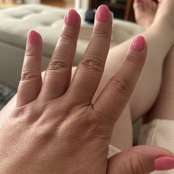 Cindy's Nails - SNS Powder Gel #192 Essie Polish #1833 Loud & Proud - A close-up of a hand with pink nail polish resting on a person's leg, with a blurred background of a living room. - Nail Salons Near Me - Harrison, New York