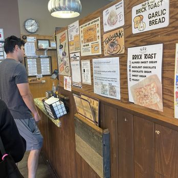 Half and Half Tea Express - A customer is standing at the counter of a bubble tea shop, facing the menu boards on a wooden wall. The menu includes various drinks such as brick toast, garlic bread, and specialty teas. The shop interior features a ceiling with a hanging light fixture, a clock, and a small window or display area in the background. The customer is dressed casually in a gray t-shirt and shorts. - Coffee & Tea Near Me - Pasadena, California