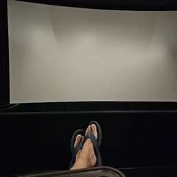 A person wearing flip-flops is seated in a movie theater, with their feet visible in front of a large, blank projection screen.
