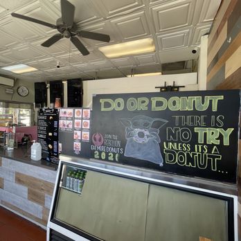 BC’s Donuts - Inside a donut shop, a chalkboard sign features a drawing of Yoda from Star Wars and humorous text about donuts, including phrases like "DO OR DONUT" and "NO TRY UNLESS IT'S A DONUT." The sign is positioned on a stand near the counter, which has a menu, a hand sanitizer bottle, and other items. The ceiling has decorative panels and a ceiling fan, and part of the shop's interior, including a clock and menu boards, is visible. - Donuts Near Me - Pasadena, California