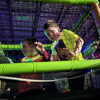 Chasers Laser Tag - Three children are inside a netted play structure at an indoor entertainment venue, surrounded by bright green padded bars and black netting. The background features purple and blue lighting, indicating a lively atmosphere suitable for activities like laser tag, arcade games, and sports. The children appear engaged and curious, with one holding a device and another looking at the camera. - Laser Tag Near Me - Naperville, Illinois