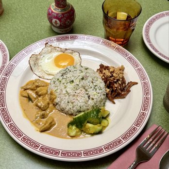 Kelang - Nasi Lemak - A round white plate with a red decorative border holds a serving of rice topped with chopped vegetables, alongside a fried egg, curry with chicken, cucumber slices, and shredded meat. The table has a green surface, with a glass of amber-colored beverage with a lemon slice, a small red and white flower vase, and a pink napkin with a fork and spoon. The setting suggests a casual dining environment, possibly at a restaurant specializing in Asian cuisine. - Salad Near Me - Brooklyn, New York