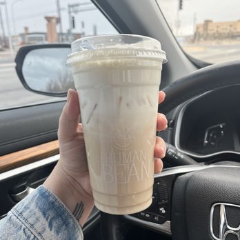 The Human Bean - Chai with bourbon cold foam - A person holds a large iced drink from The Human Bean, with the logo visible, inside a vehicle with a view of the road outside. - Coffee & Tea Near Me - Albuquerque, New Mexico