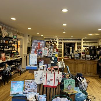 Lighthouse Keeper's Pantry - Inside - The image shows the interior of a cafe and gift shop with wooden flooring and white shelving units filled with various products. In the foreground, display tables hold bags, books, and decorative items, including a book titled 'SMUG SEAGULL' and a bag with a seagull illustration. Staff members are visible behind the wooden counter, which has a menu board and coffee-making equipment. The space is well-lit with ceiling lights, and the overall setting appears cozy and organized. - Specialty Food Near Me - Yarmouth, Massachusetts