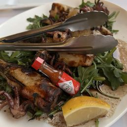 Grilled Calamari - A seafood dish featuring grilled octopus and other seafood, garnished with greens, lemon, and hot sauce, served on a white plate.