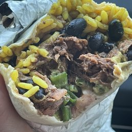 Yellow Rice - A partially eaten burrito filled with yellow rice, black beans, shredded meat, and chopped green vegetables, wrapped in foil and paper. The image shows the cross-section of the burrito, revealing the ingredients inside. The background is blurred, focusing on the food item.