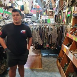 He got a new belt and pouch set - A person is inside a military surplus store with racks of military jackets, camouflage clothing, and accessories visible around.