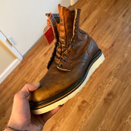 Traction tread - A person is holding a brown leather boot with a white rubber sole indoors on a wooden floor. The boot shows signs of wear, with creases and scuffs on the leather. It has metal eyelets for laces, but no laces are present. The interior lining is visible at the top, and a red tag is attached to the boot. The person's hand is visible, wearing a bracelet, and the person's shoes are seen at the bottom of the image.