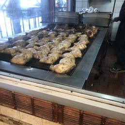 Chicken being cooked - Chicken pieces are arranged on a large grill inside a restaurant, with some pieces appearing partially cooked. The grill is situated near a window, allowing natural light to illuminate the scene. In the background, a person is seen preparing food, with kitchen equipment and tiled walls visible. The restaurant has a brick and tile exterior and appears to serve grilled chicken dishes.