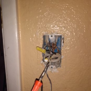 Lowe's Home Improvement - The inside of my light switch - A wall with a beige textured surface features an electrical outlet box with no cover plate. Several wires, including a black wire with a red connector and other unconnected wires, are visible, indicating ongoing electrical work or repair. The surrounding wall appears intact, but the outlet is incomplete and exposed. - Home Decor Near Me - Phoenix, Arizona