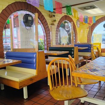 El Coyote - The photo depicts the interior of a Mexican restaurant with booths along a wall featuring brick arches and large windows. The booths have wooden frames with blue, green, and yellow upholstery. Colorful papel picado banners hang from the ceiling, adding decorative flair. The floor is tiled with reddish-brown tiles, and there are wooden chairs and tables with tiled surfaces. The windows allow natural light to illuminate the space, and a speaker is mounted on the wall near the ceiling. - Mexican Near Me - Alsip, Illinois