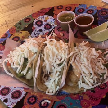 Little Joe's - Shrimp tacos - Three tacos topped with shredded cheese and other ingredients are served on a colorful plate with lime wedges and small cups of salsa and green sauce. - Cocktail Bars Near Me - Chicago, Illinois