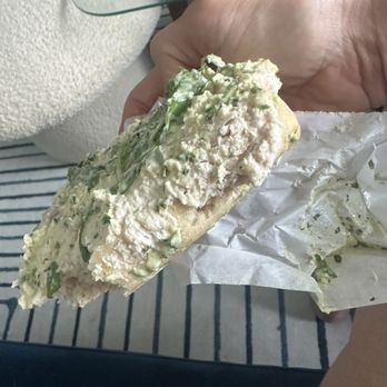 Meat & Bread - Side view of bread with thinnest layer of mayo and a little pinch of chicken. No chicken in the chicken salad. - A person holds a sandwich with a creamy filling and chopped green herbs, wrapped in paper, with a background of a striped rug and furniture. - Sandwiches Near Me - Austin, Texas