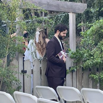Savage Suiting - Groom's suit - A man in a dark suit holding a wrapped gift stands outdoors near a wooden fence and lush greenery, with chairs in the foreground. - Formal Wear Near Me - Placentia, California