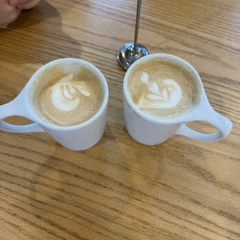 Southpine Bakery - Vanilla latte's - Two white ceramic coffee mugs with latte art sit on a wooden table, with a metal table marker in the background. - Coffee & Tea Near Me - Eugene, Oregon
