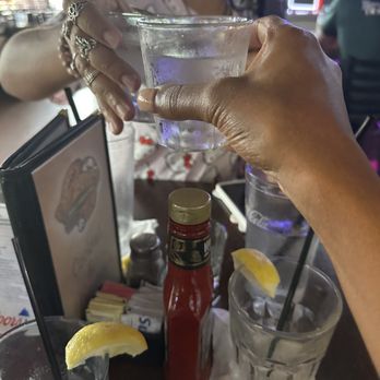 Fat Willy’s - Two people are raising glasses of water with lemon slices in a restaurant or bar setting. One person’s hand is visible, holding a glass, while the other person’s face is partially visible in the background. The table has condiments, a menu, and a napkin. The environment includes other patrons, a menu, and neon signs in the background, indicating a casual dining or bar atmosphere. - Sports Bars Near Me - Gilbert, Arizona