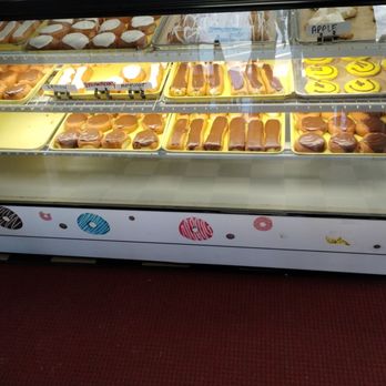Steve's Bakery - A glass display case showcases an assortment of donuts and pastries arranged on yellow trays. The selection includes chocolate-covered donuts, filled donuts, and decorated cookies with smiley faces. Small labels indicate flavors such as lemon, strawberry, apple, and cherry. The case is decorated with a colorful, donut-themed design on the lower panel, and the background shows additional baked goods and labels. - Bakeries Near Me - Ottawa, Illinois