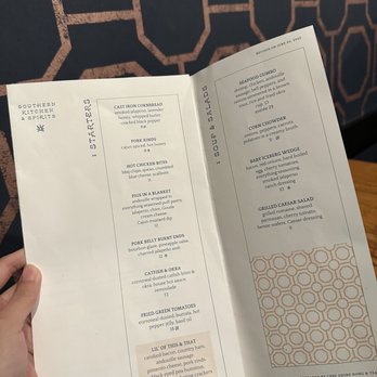 Subourbon - The menu from Southern Kitchen & Spirits displays a list of starters and sour & salads. The starters include cast iron cornbread, pork rinds, hot chicken bites, pigs in a blanket, pork belly burnt ends, catfish & okra, fried green tomatoes, and Lil' of This & That. The sour & salads section features seafood gumbo, corn chowder, baby iceberg wedge, and grilled Caesar salad. The menu is printed on white paper with black text and is held by a person in front of a patterned wall. - Southern Near Me - Columbus, Ohio