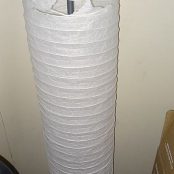Prohands Movers - Tore up my lamp shade and I don't know why that was even put on the truck. - A tall, cylindrical white paper lantern with a tear near the top section, leaning against a plain, light-colored wall. The lantern appears to be made of thin paper with horizontal ridges and is positioned on the floor, with some boxes and objects nearby. - Movers Near Me