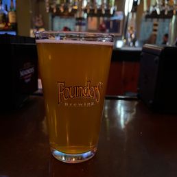 Chilling at the bar - A pint glass filled with beer featuring the Founders Brewing logo is on a bar counter, with a blurred bar background including taps and bottles.