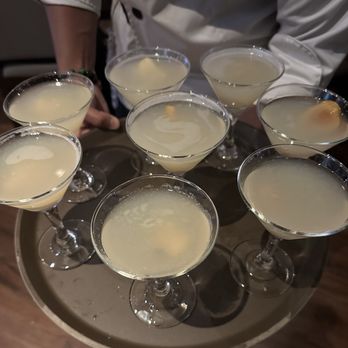 Bonefish Grill - A person dressed in a white chef's coat is holding a tray with eight cocktails in martini glasses. The drinks are pale yellow or light-colored, with some garnished with fruit. The background is blurred, focusing on the tray and drinks. - Seafood Near Me - Gambrills, Maryland