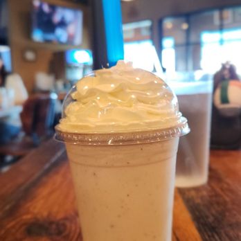 Mac's Local Eats - Carmel crunch milkshake - A caramel crunch milkshake with whipped cream and a blue straw is placed on a wooden table inside a restaurant with a lively interior. - American Near Me - St Louis, Missouri