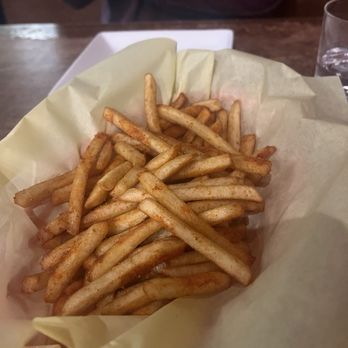 310 Eatery - House Fries - A basket filled with seasoned French fries is placed on a table, lined with yellow parchment paper. The fries are golden-brown and appear crispy, with some seasoning visible. In the background, part of a person's arm and a glass of water are visible, indicating a dining setting. - Burgers Near Me - Albany, California