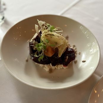 Haven Restaurant and Bar - Smoked Beets - A dish of smoked beets is served in a white bowl, garnished with microgreens, cheese, and small vegetable pieces. The presentation is simple, with the ingredients arranged on a bed of crumbled elements, and the bowl is placed on a white tablecloth. The background shows a blurred table setting. - Southern Near Me - Atlanta, Georgia