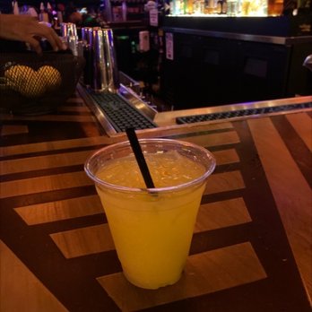 Cobra Arcade Bar - Tito's screwdriver - A yellow beverage served in a clear plastic cup with a black straw, positioned on a wooden bar counter with a striped pattern. The background shows bar equipment, bottles, and a dimly lit bar environment. - Bars Near Me - Phoenix, Arizona