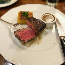 Filet, medium rare - A plate features a medium-rare filet mignon with a pink interior, a small serving of potato gratin topped with herbs, and a cup of sauce, served at a restaurant.