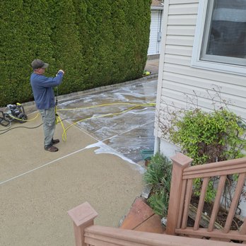 VB Renovations - Powerwashing - A person is engaged in power washing a concrete patio adjacent to a house with white siding. The individual is holding a power washer wand, with foam and water on the surface of the patio. Nearby, a power washer machine is connected to the wand via a yellow hose. The scene includes a large, well-maintained green hedge in the background and part of a wooden staircase with potted plants in the foreground. The sky is partly cloudy, indicating a clear day. - General Contractors Near Me